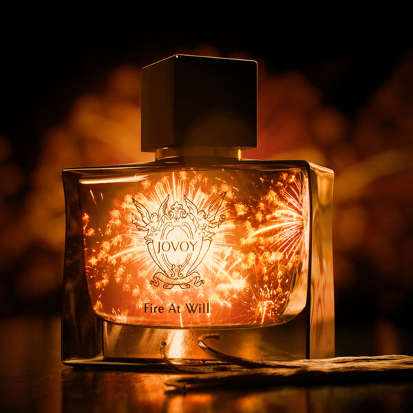Jovoy - Fire at Will Parfumflakon in warmem, goldenem Licht mit Feuerwerk im Hintergrund vor dunklem Ambiente