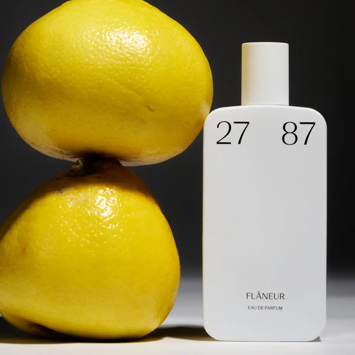 27 87 Perfumes - flâneur Parfumflakon in schlichtem weiß neben zwei frisch aussehenden Zitronen vor dunklem Hintergrund